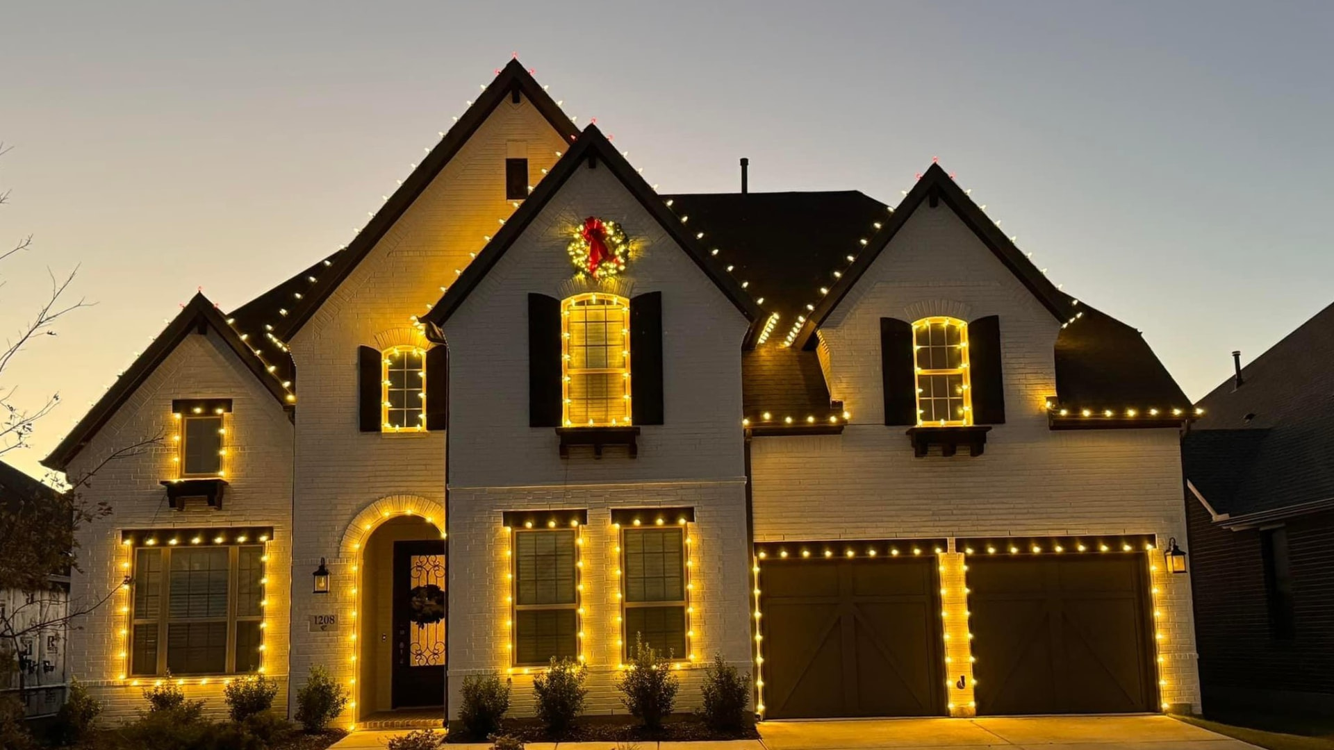 Two-story brick home in the Dallas-Fort Worth area at dusk, with warm white Christmas lights outlining every roofline, window and garage door, and a lit wreath on the front gable