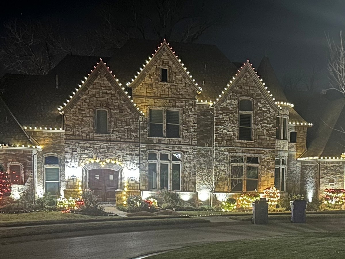 Large two-story stone home with steep gables outlined in warm white lights, with lit garland along the front beds